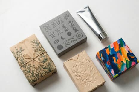 Fünf Produktverpackungen im Flatlay zeigen die Packaging-Design-Trends 2026: botanische Illustration auf Kraftpapier, minimalistischer Graukarton mit Blockschrift, silberne Metalltube, haptisch geprägte Naturfaser-Verpackung und farbintensive Artist-Showcase-Box mit expressiven Pinselstrichen.